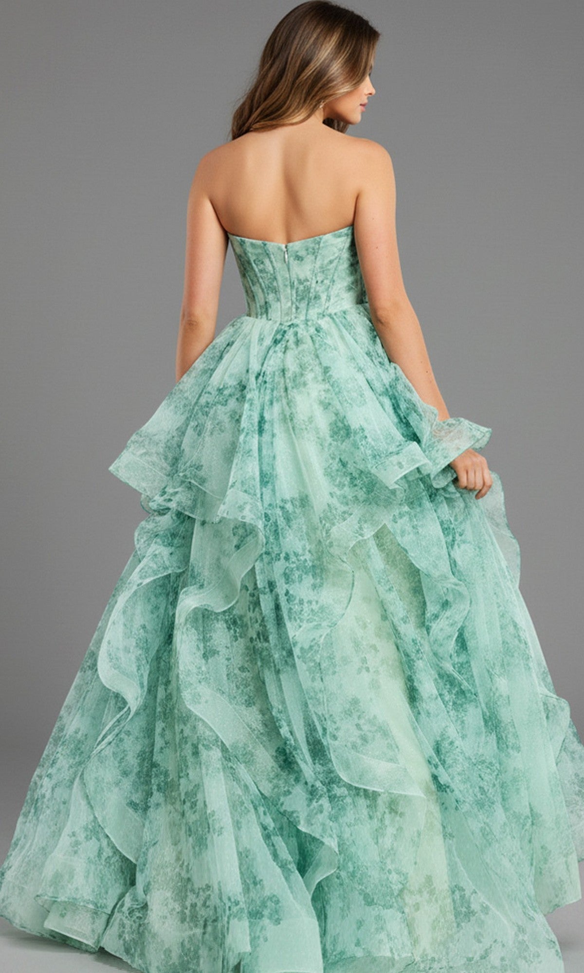 A woman faces away from the camera in a Jovani 49777 Strapless Green Watercolor Prom Gown. The long A-line dress features layered mint green fabric with a delicate floral pattern, all set against a gray background for an elegant look.